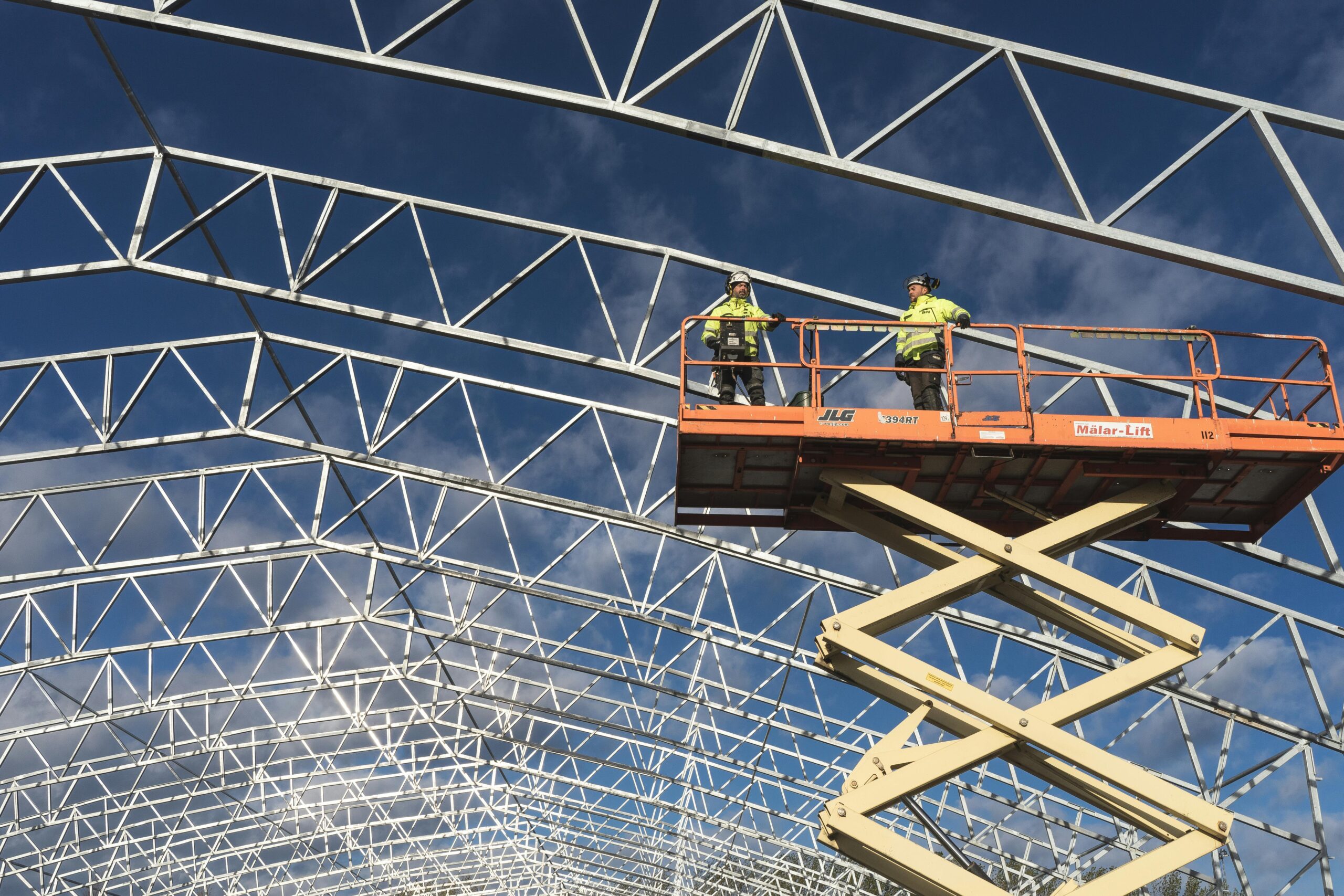 Two workers on a scissor lift at a construction site, framed by a metal structure against a blue sky.