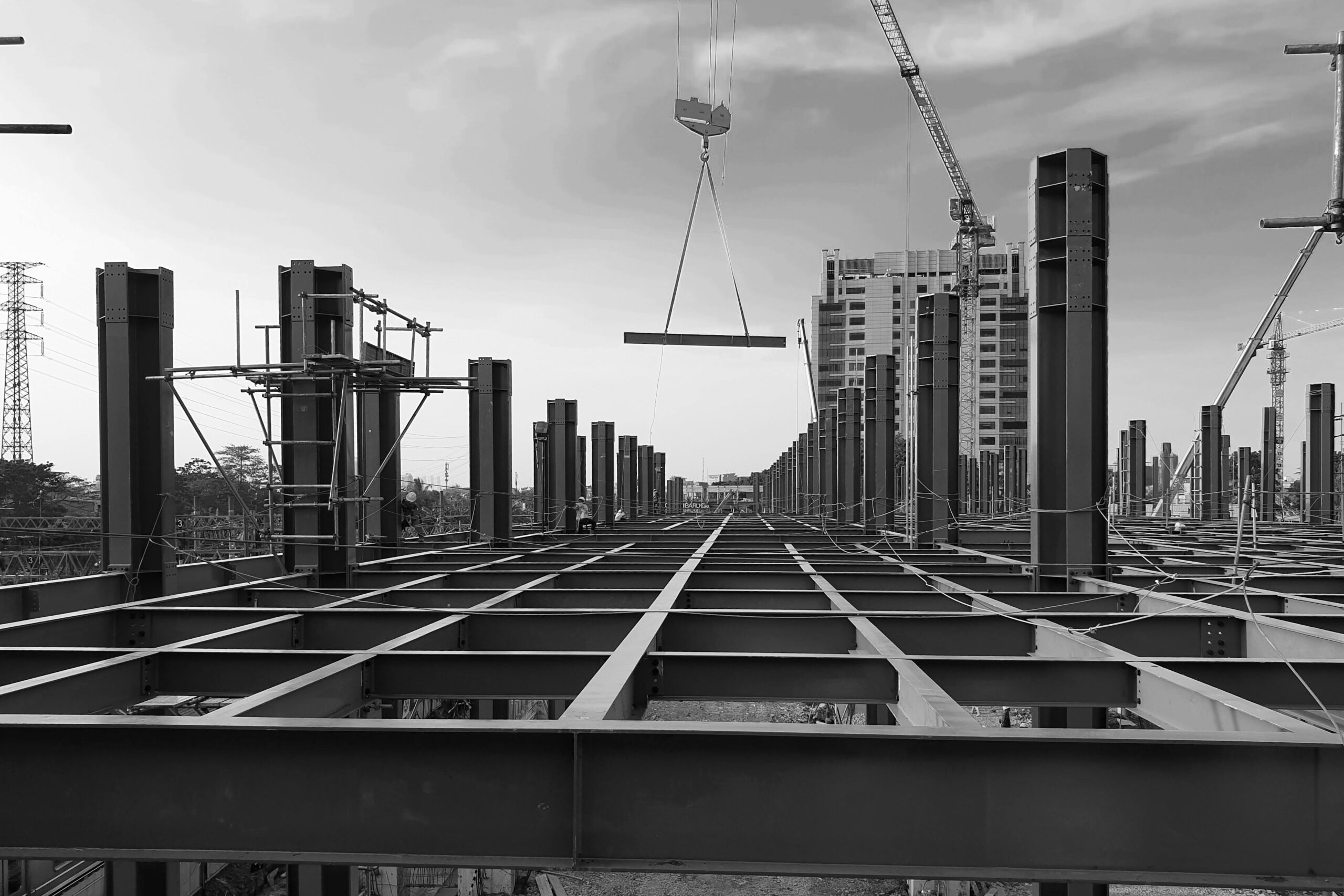 Black and white view of a modern urban construction site in Indonesia with steel beams and cranes against a cloudy sky.